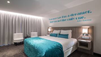 Ein modernes Hotelzimmer mit einem großen Bett, zwei Nachttischen, einem Zitat an der Wand und Blick auf die Stadt durch das Fenster.
