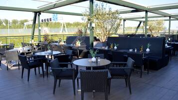 An outdoor seating area with round tables and chairs, a covered section with a sofa, and a view of the surrounding landscape.