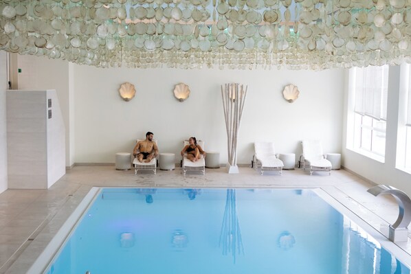 An indoor swimming pool with a clear blue water surface, surrounded by white lounge chairs and a decorative ceiling.