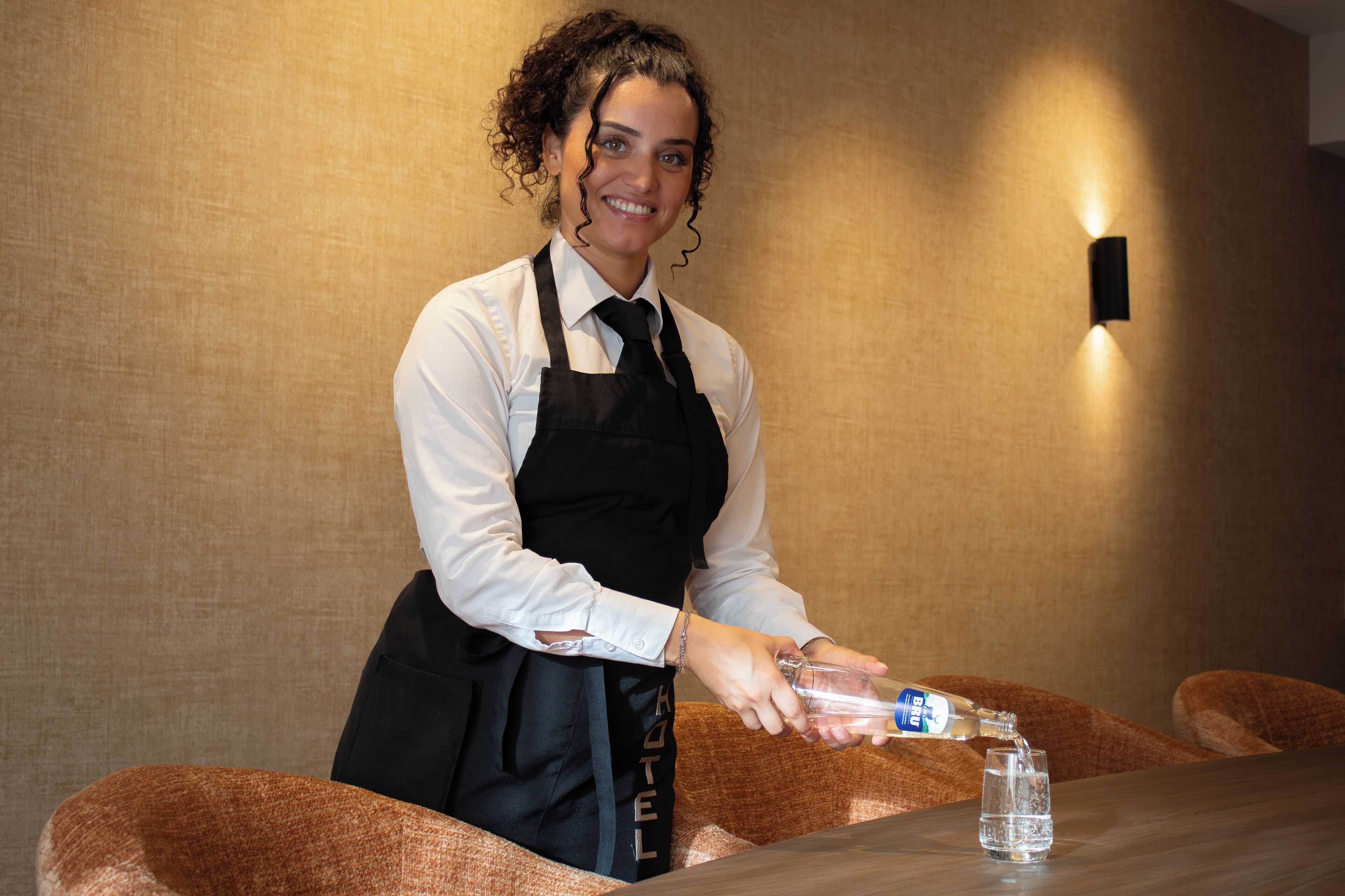 A person pouring a drink into a glass on a table in a room with a textured wall and a wall-mounted light fixture.