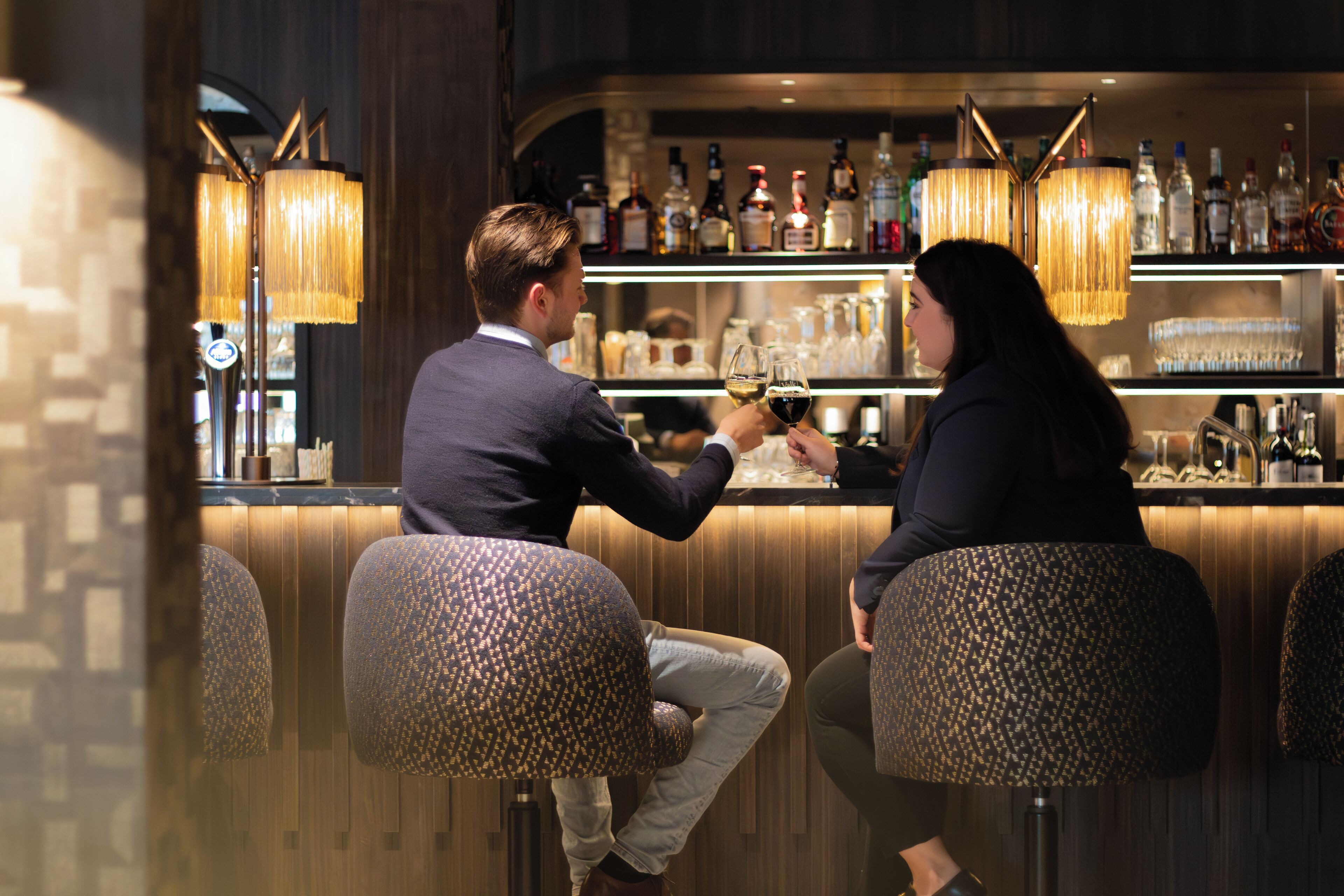 A bar area with two stools, a bar counter with various bottles, and hanging lamps.