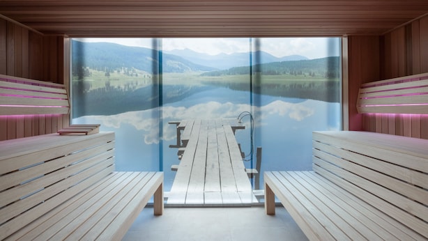 A sauna room with wooden benches and a large window offering a view of a lake and mountains.