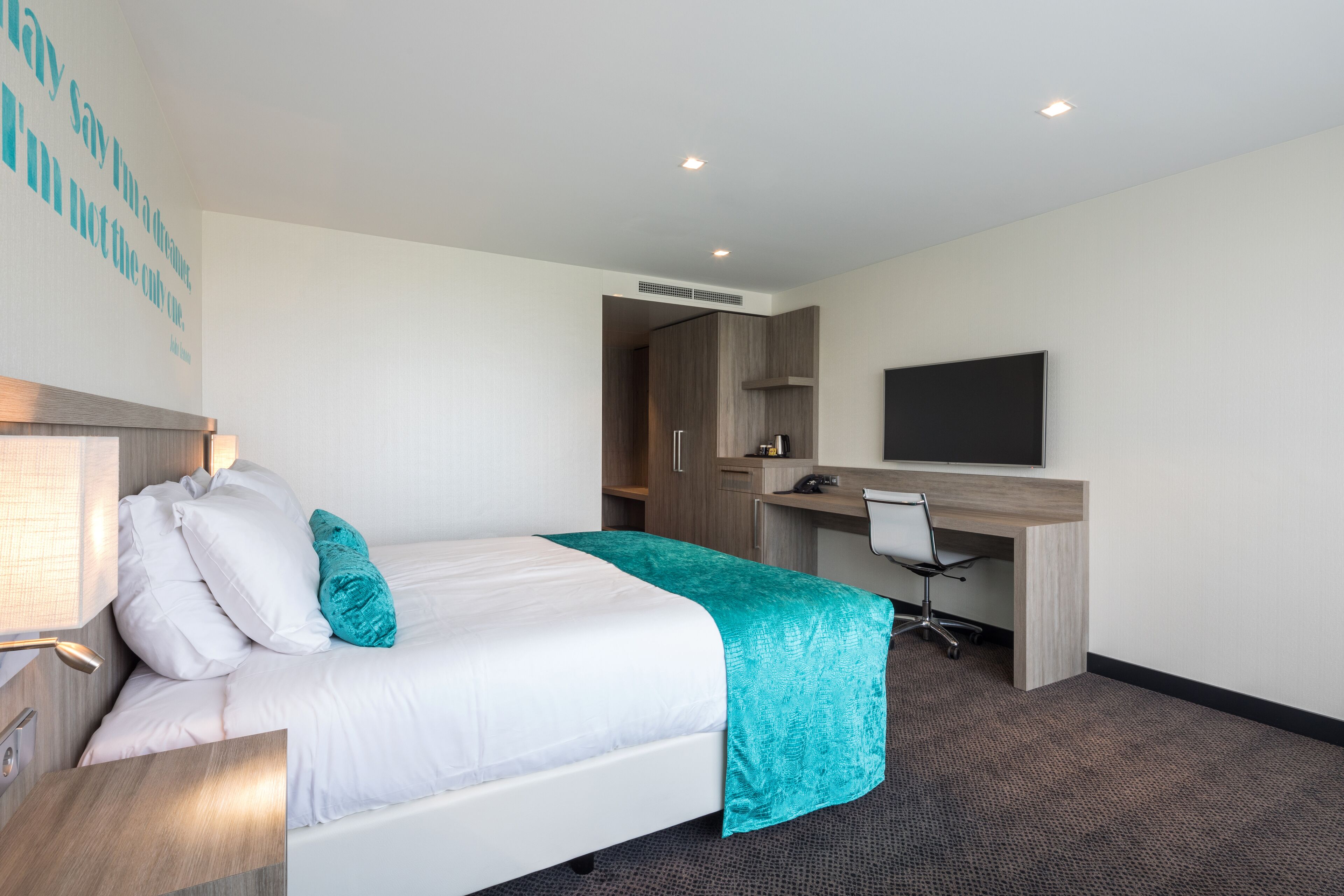 Une chambre d’hôtel équipée d’un lit, d’un bureau, d’une chaise, d’une télévision et d’un mur orné d’une citation.