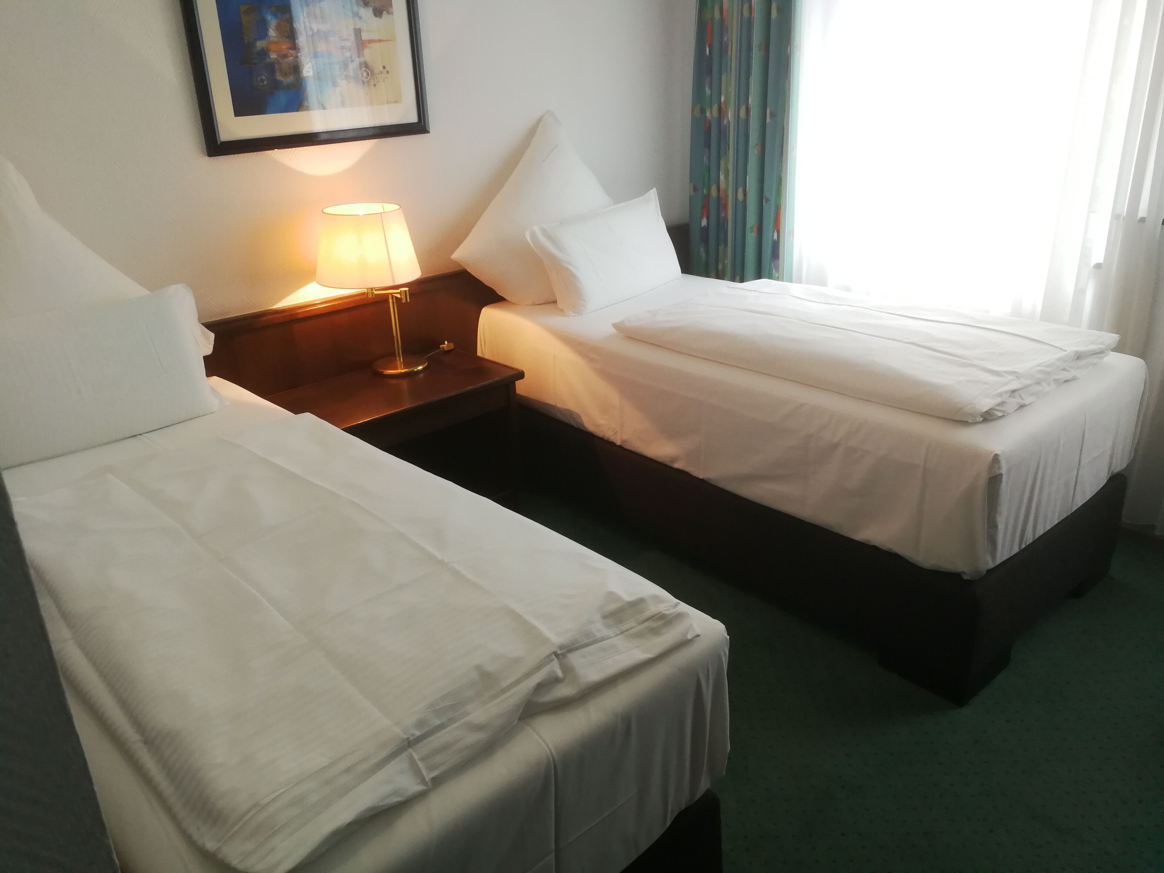 A hotel room with two single beds, a nightstand with a lamp, a framed picture on the wall, and a window with curtains.