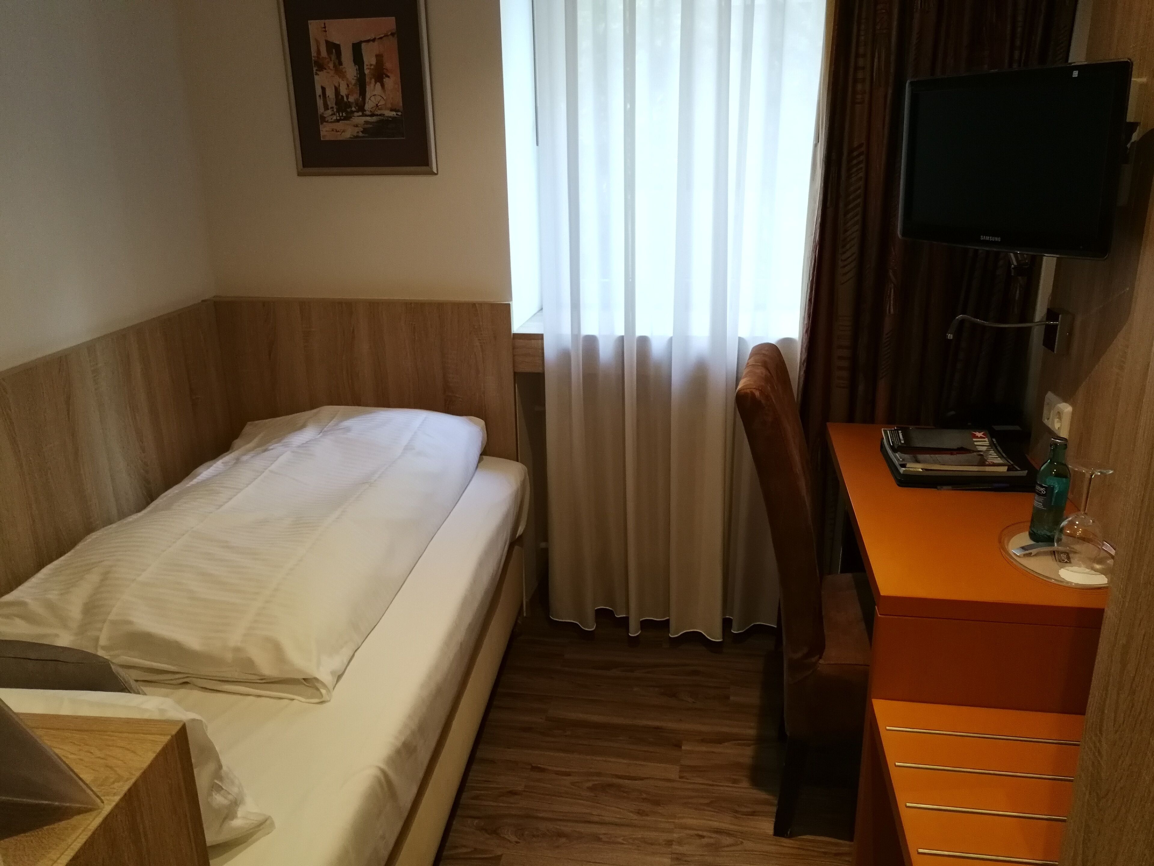 A hotel room with a single bed, a wooden headboard, a desk with a chair, a television, and a window with curtains.