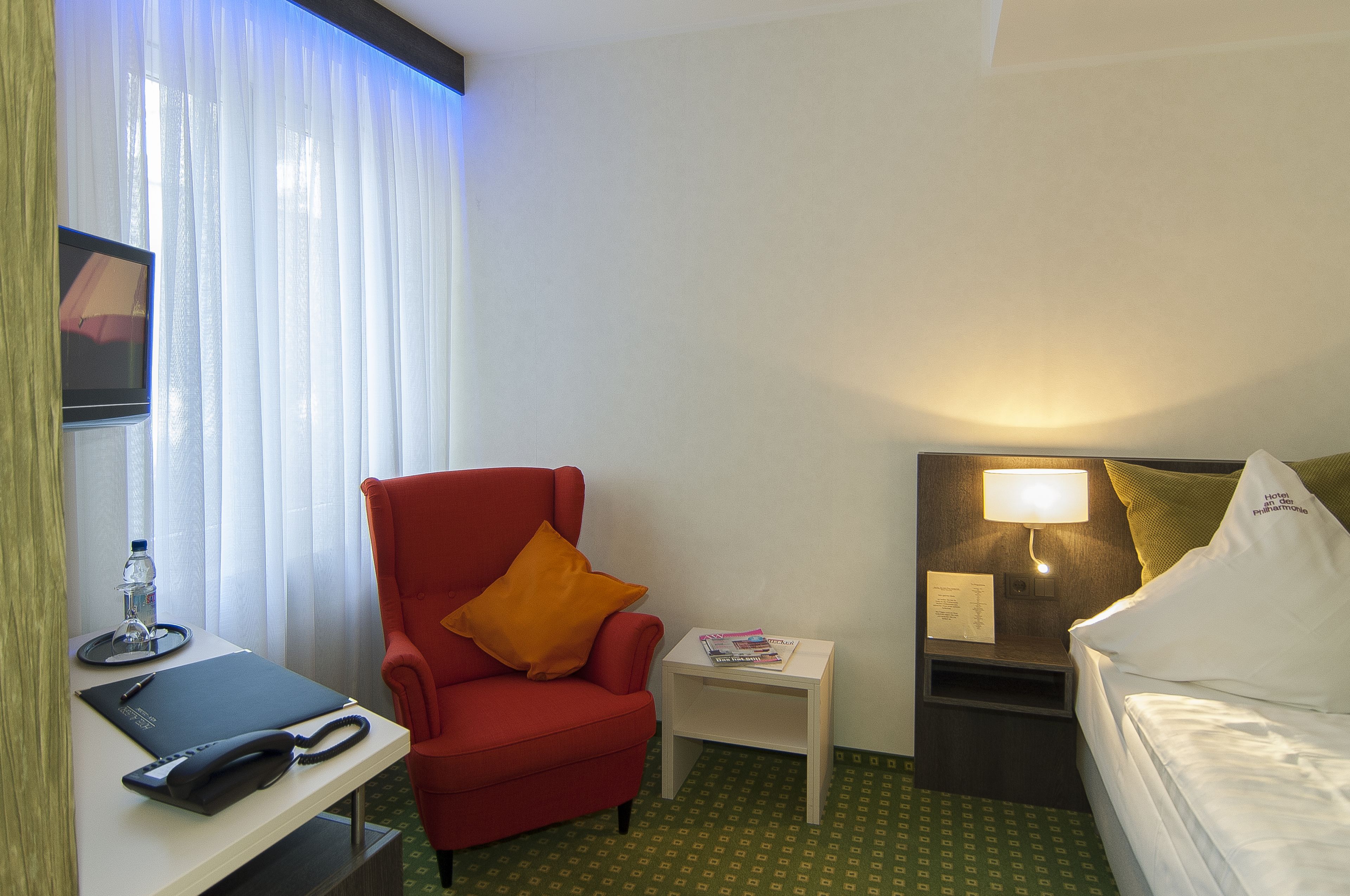 A hotel room with a red armchair, a small bedside table with a lamp, a bed with white bedding, and a desk with a chair and a water bottle.