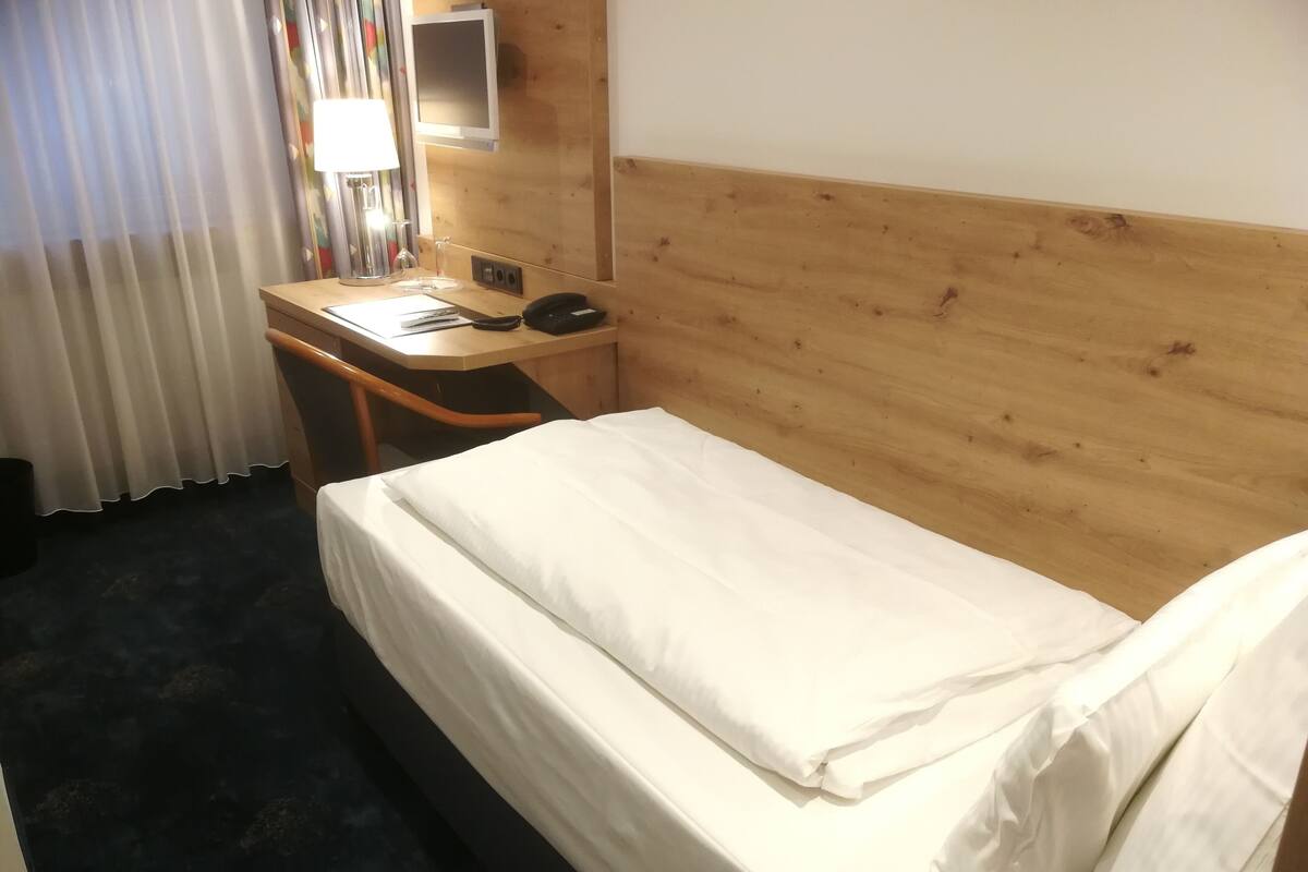 A hotel room with a wooden headboard, a desk with a lamp, a phone, and a bed with white linens.