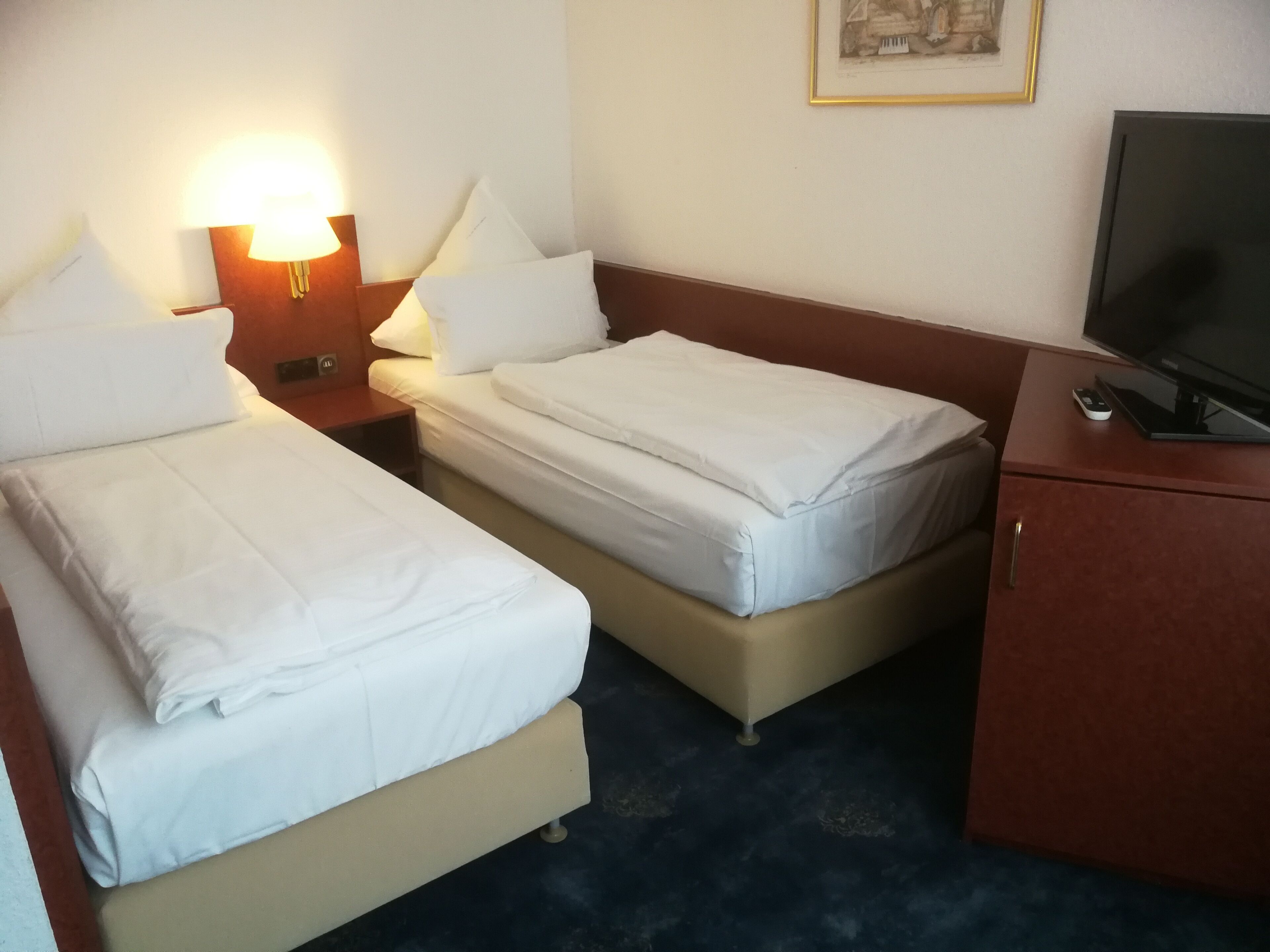 Hotellihuone, jossa on kaksi sänkyä, yöpöytä lampun kanssa, televisio, joka on sijoitettu kaappiin, ja seinällä oleva kehystetty kuva.