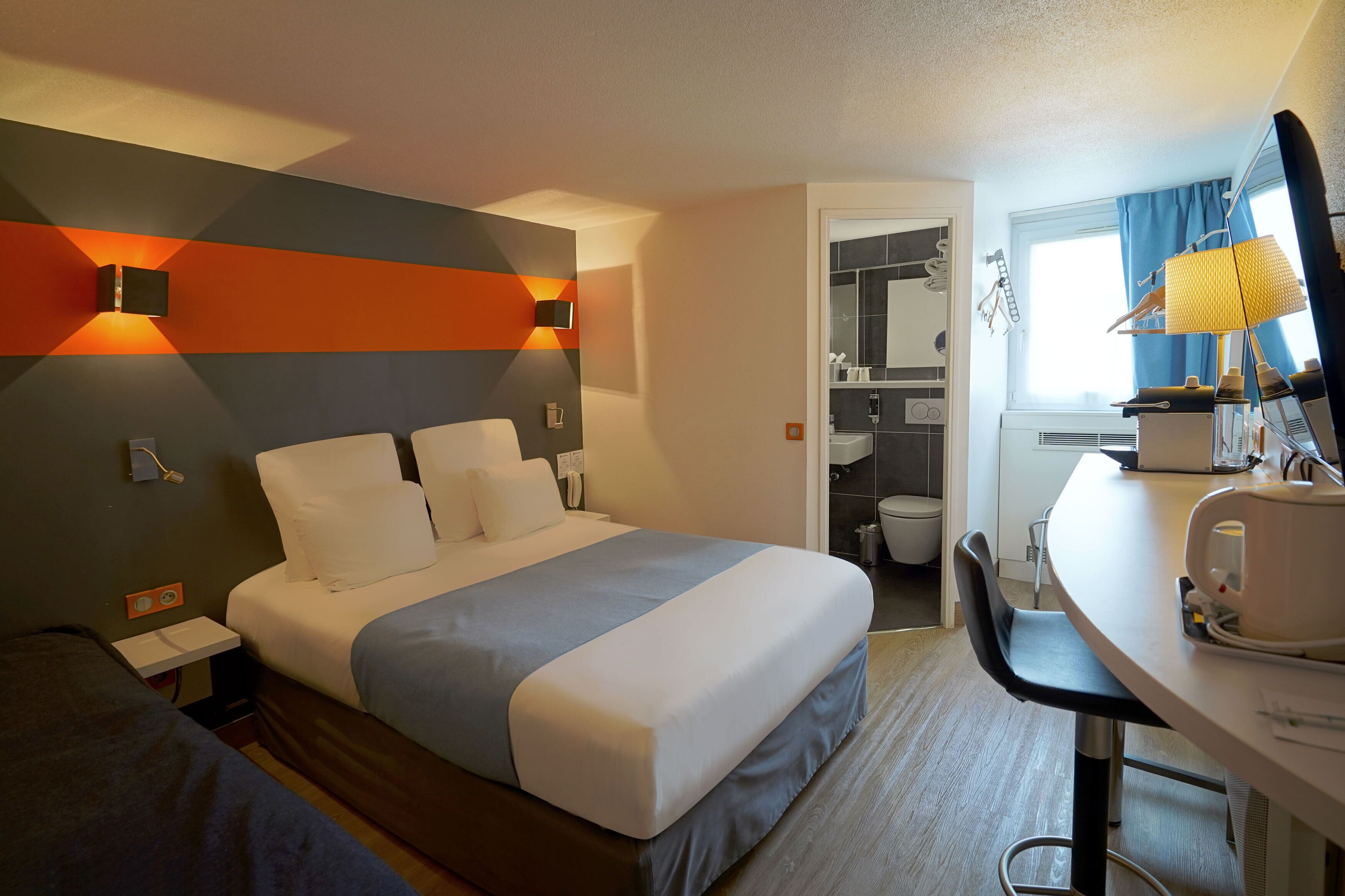 Une chambre d’hôtel avec un lit, une table de chevet et un bureau avec une lampe. On aperçoit une salle de bain au fond.