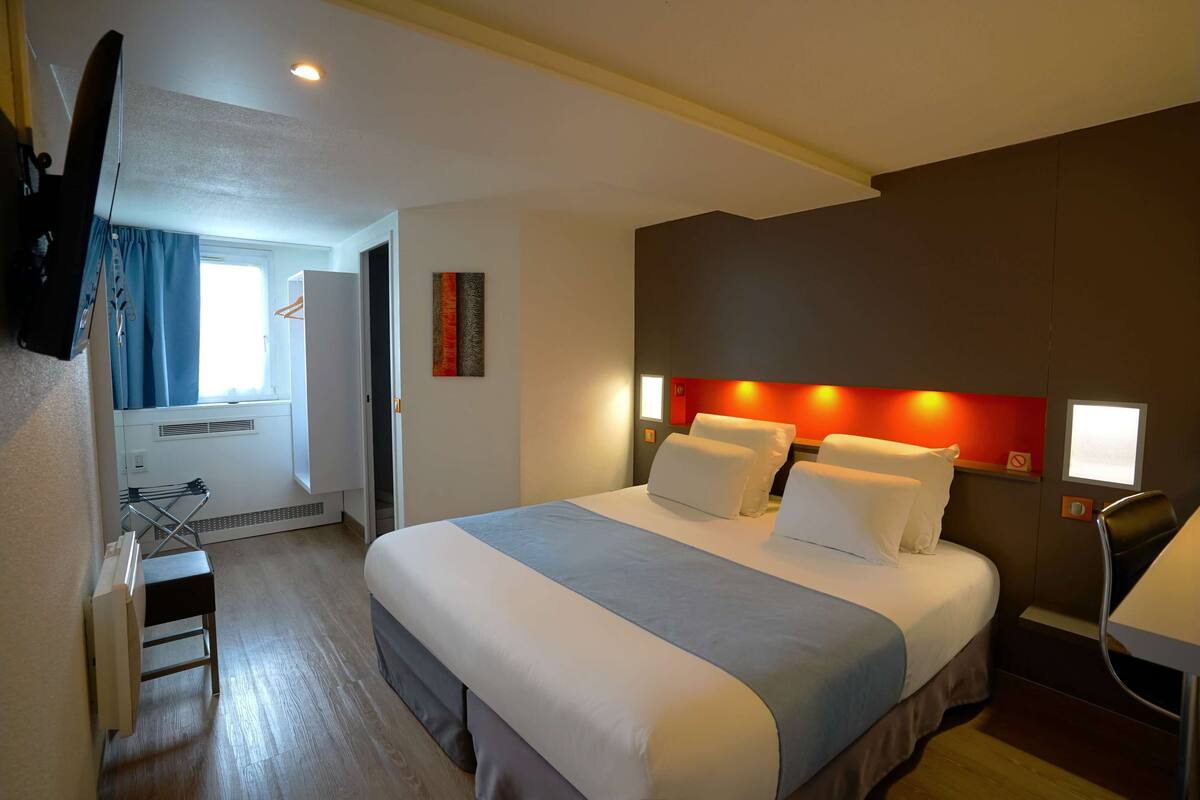 Ein Hotelzimmer mit einem großen Bett, einem Schreibtisch, einem Stuhl, einem an der Wand befestigten Flachbildfernseher und einem Fenster mit Vorhängen.