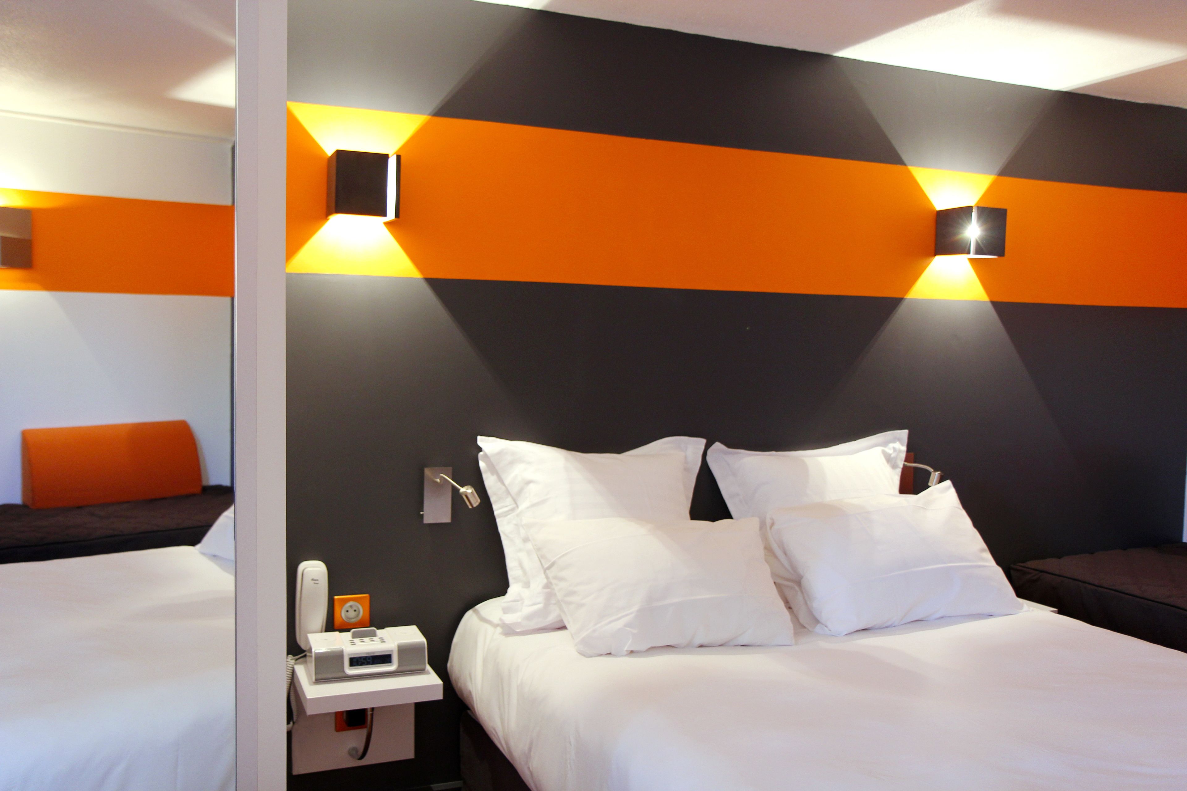 A hotel room with a bed, white bedding, a bedside table with a phone and a small orange device, and two wall-mounted light fixtures.