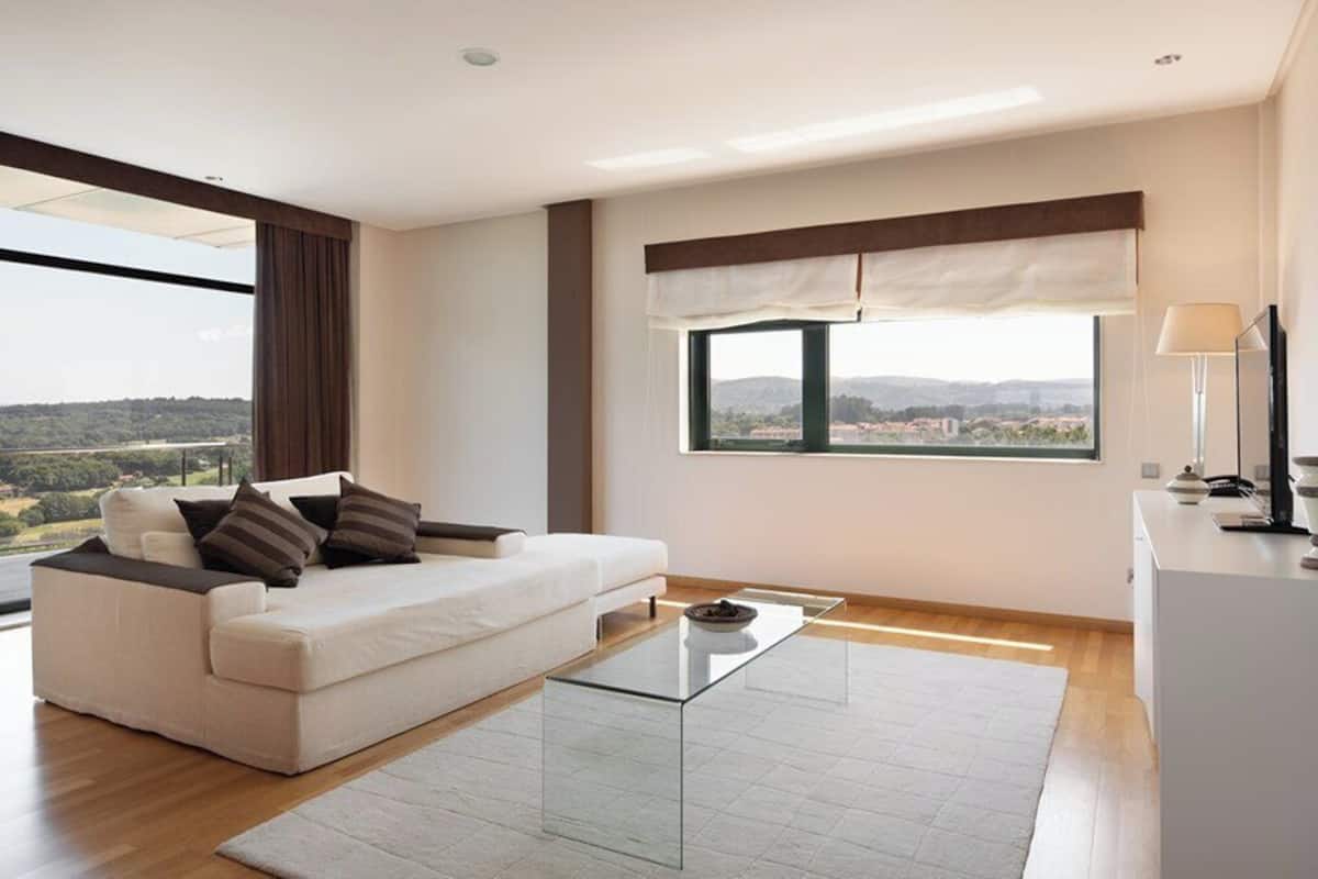 A modern living room with a white sofa, a glass coffee table, and a large window offering a view of the countryside.