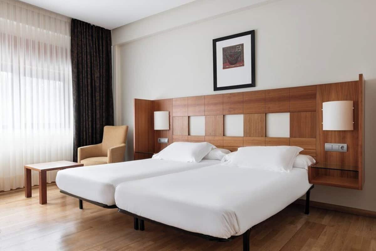 A hotel room with two beds, a chair, a small table, and a framed picture on the wall.