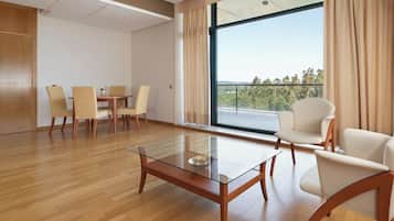 Quarto de hotel moderno com uma mesa de centro de vidro, área de refeições com cadeiras e uma varanda com vista para árvores.