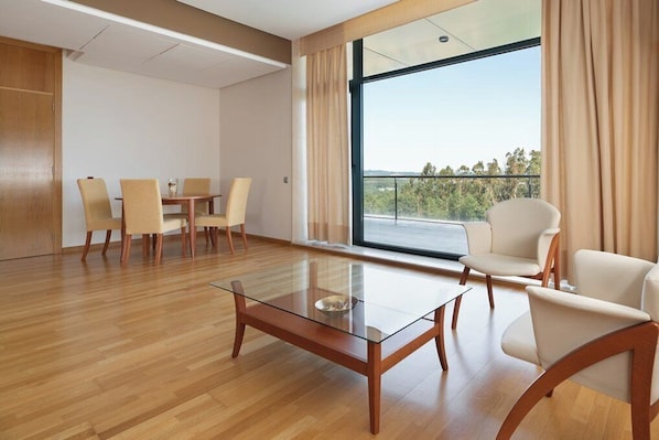 Una habitación de hotel moderna con una mesa de centro de vidrio, un área de comedor con sillas y un balcón con vistas a árboles.