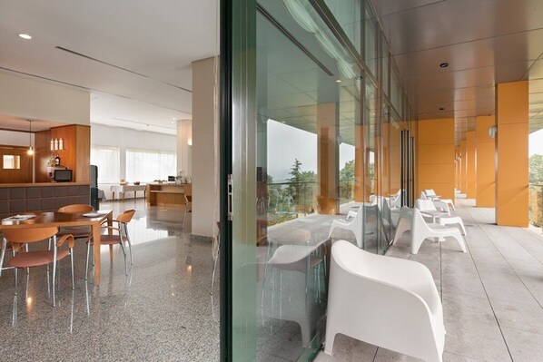 A modern hotel lobby with a glass partition, a dining area with wooden tables and chairs, and a seating area with white plastic chairs. - Exe Via Argentum (Silleda)