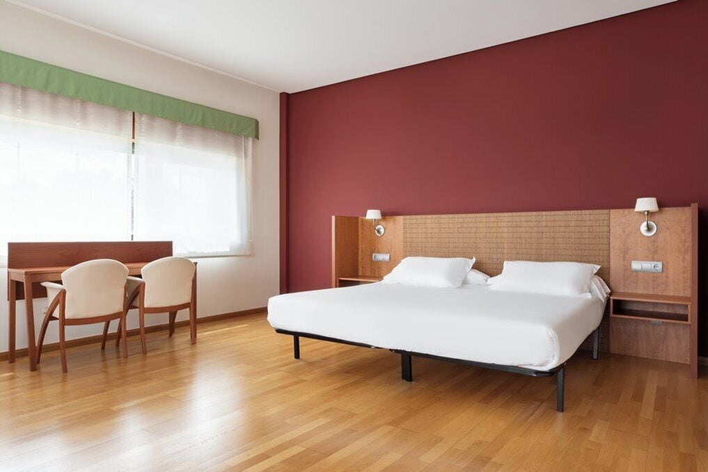 A hotel room with a wooden floor, a bed with white linens, a desk with chairs, and a window with a green curtain.