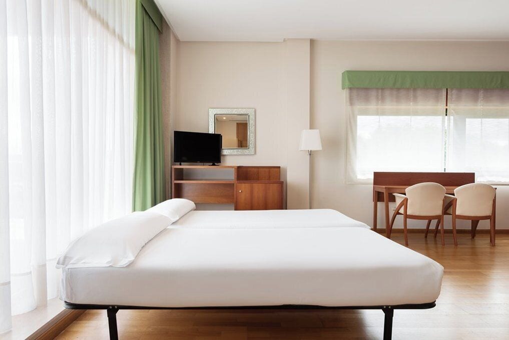 Une chambre d’hôtel avec un lit simple, un bureau avec une chaise, une télévision et une fenêtre avec des rideaux légers.