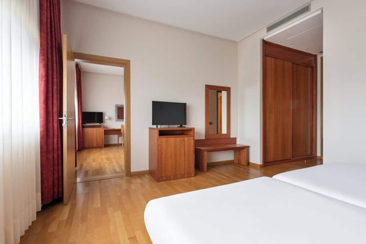 A hotel room with a wooden floor, a bed, a TV, a desk, a mirror, and a wardrobe.
