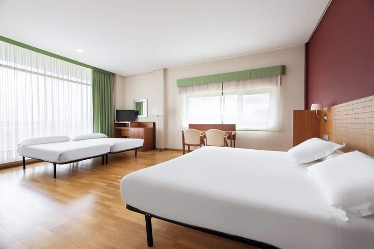 A hotel room with a large bed, two single beds, a desk with a chair, a TV, and a window with curtains.
