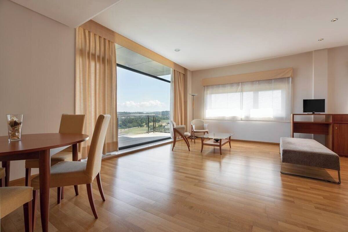 A modern hotel room with a large window offering a scenic view, a dining area with a table and chairs, a sofa, and a flat-screen TV.