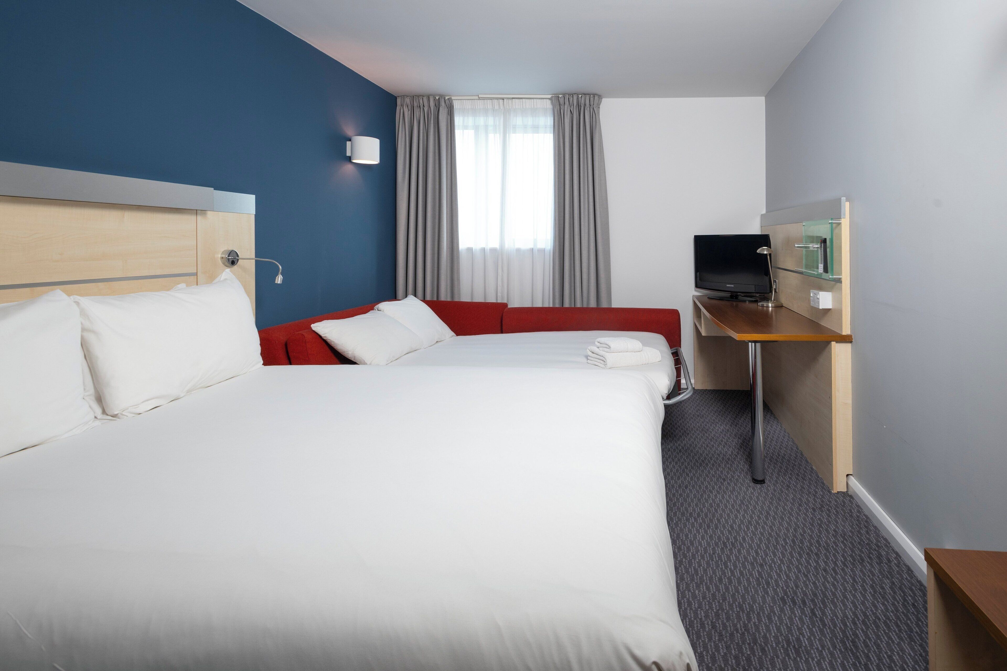 Una habitación de hotel con cama, escritorio, televisor y ventana con cortinas.