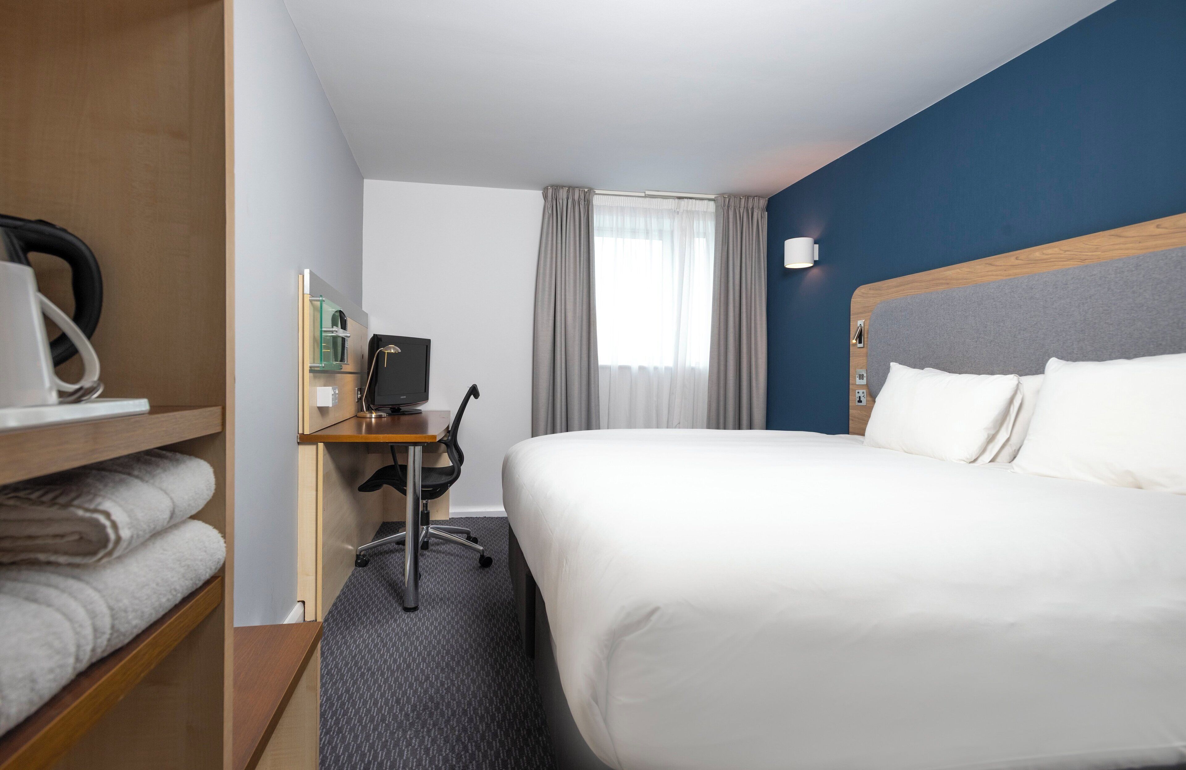 A hotel room with a large bed, a desk with a computer, a coffee machine, and a shelf with towels.