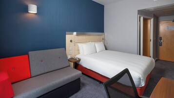 A hotel room with a bed, a gray sofa with red cushions, a small table, and a chair.