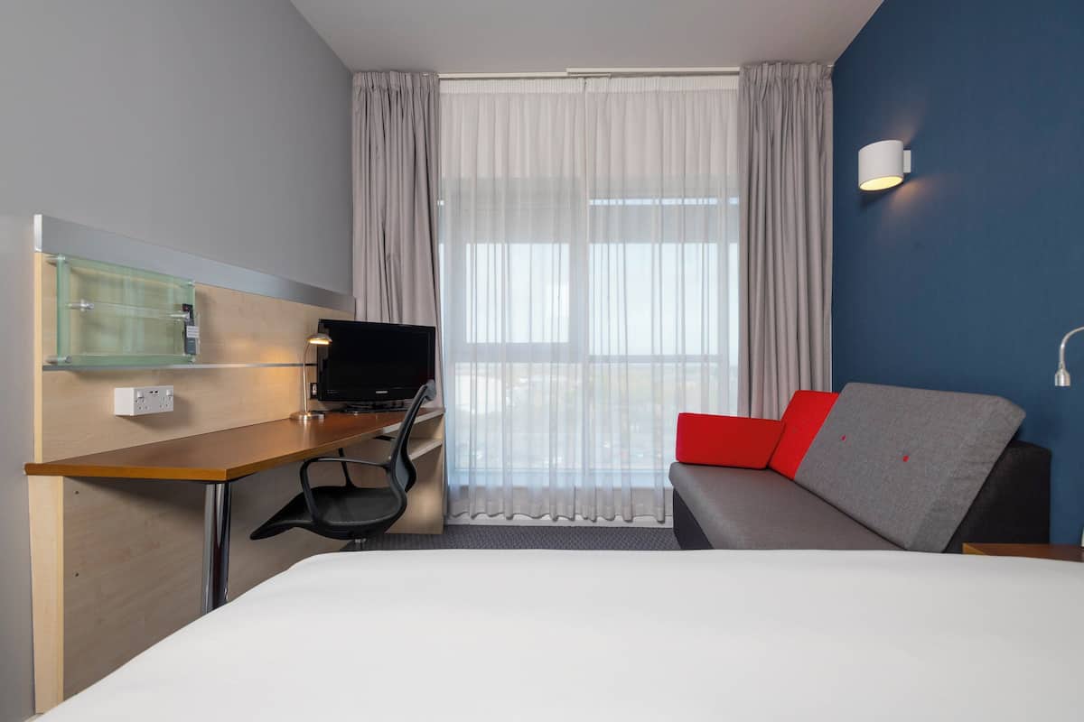 A hotel room with a bed, a desk with a computer, a chair, a sofa with red cushions, a window with sheer curtains, and a wall-mounted light fixture.