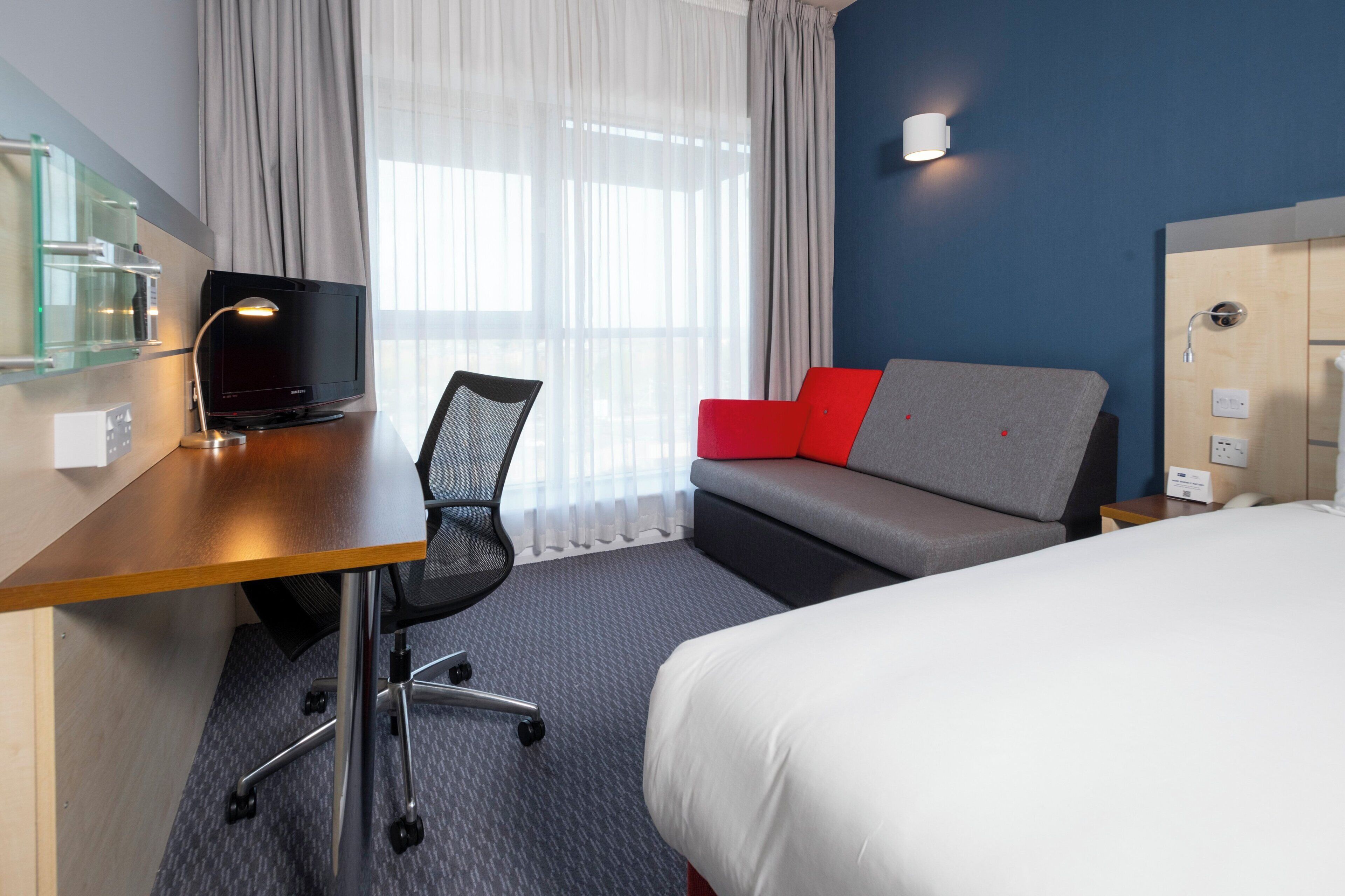 Habitación de hotel con cama, escritorio con computadora, silla, sofá con cojines rojos y zona de ducha.