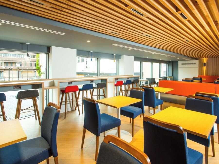 A modern dining area with wooden ceiling slats, multiple seating arrangements, and large windows offering views of the outside.