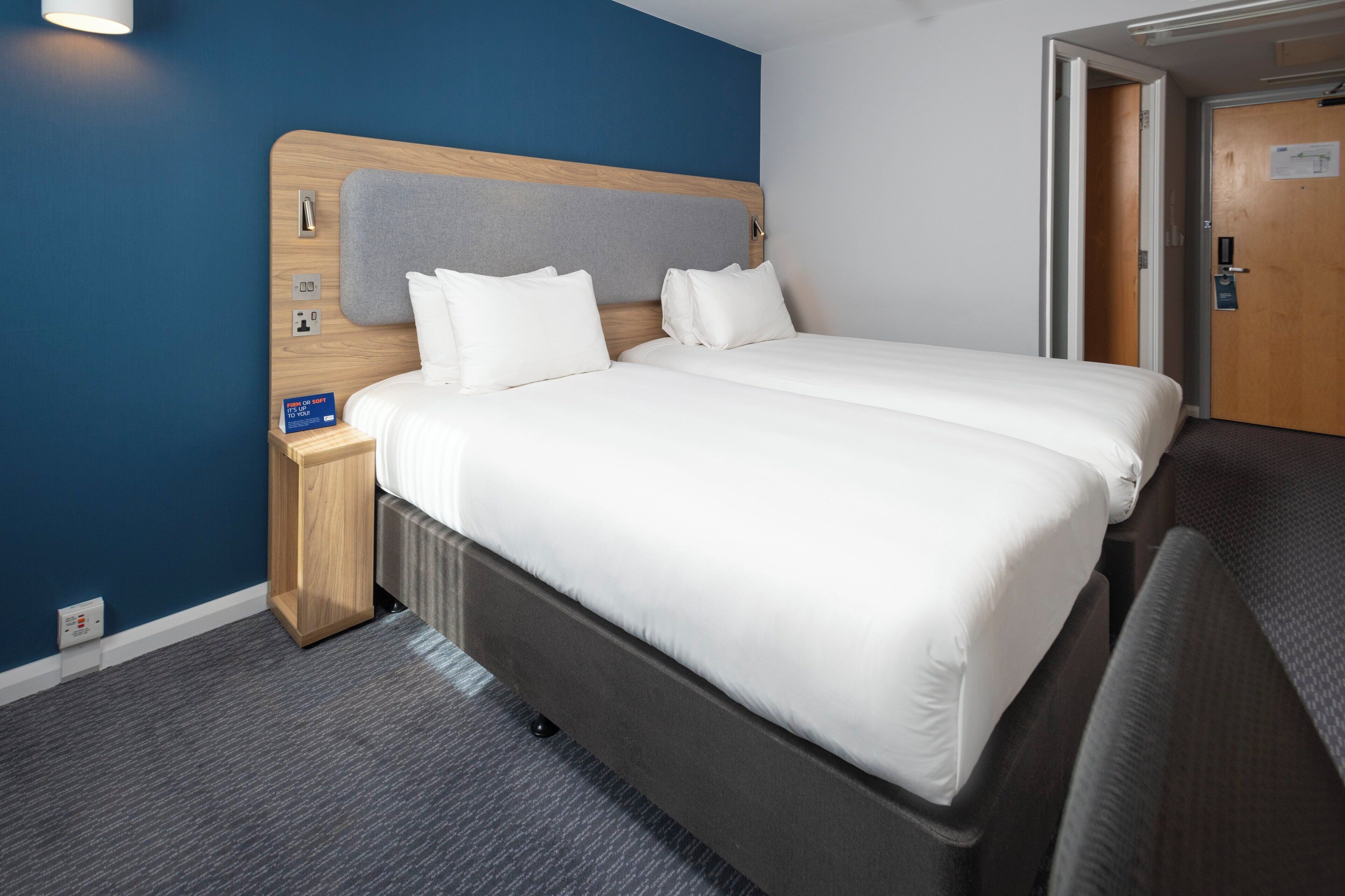 A hotel room with a double bed, a wooden headboard, a nightstand, and a door to the right.