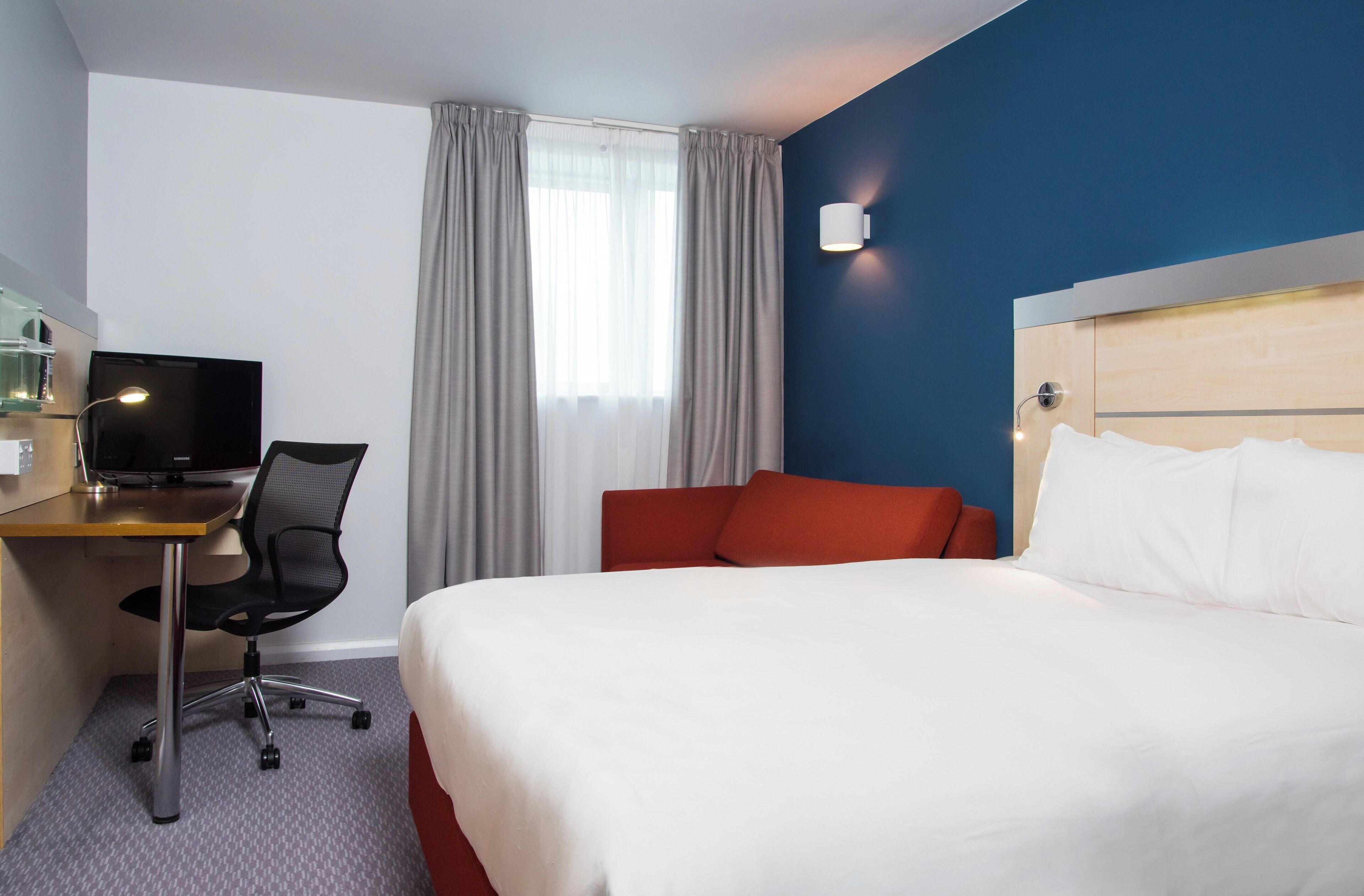 Habitación de hotel con cama, escritorio con ordenador, silla, televisor y ventana con cortinas.
