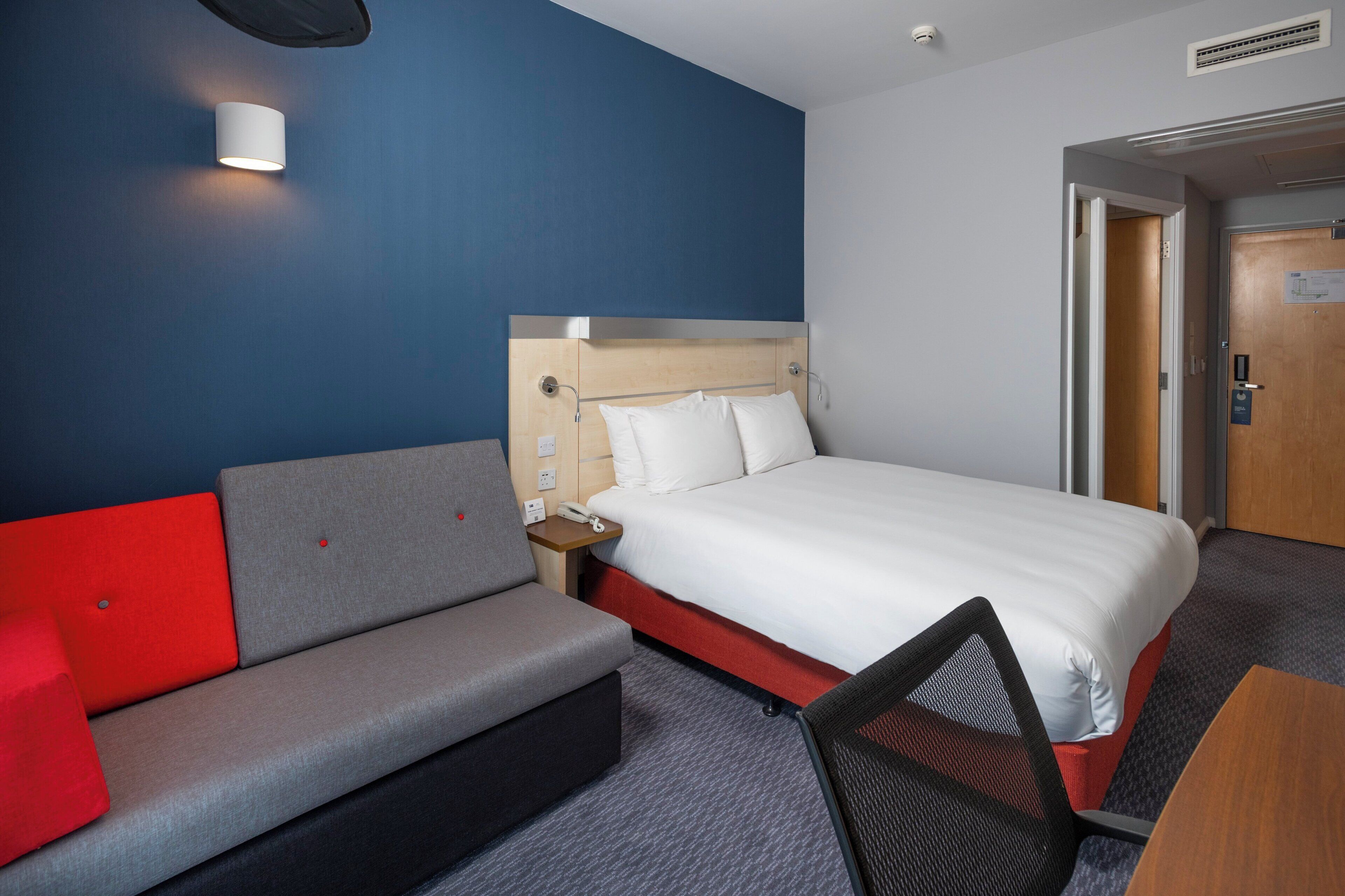 Habitació d'hotel amb un llit, un sofà vermell i gris, una taula petita i una cadira.