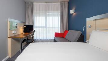 Habitación de hotel con cama, escritorio con computadora, silla, sofá con cojines rojos y ventana con cortinas.