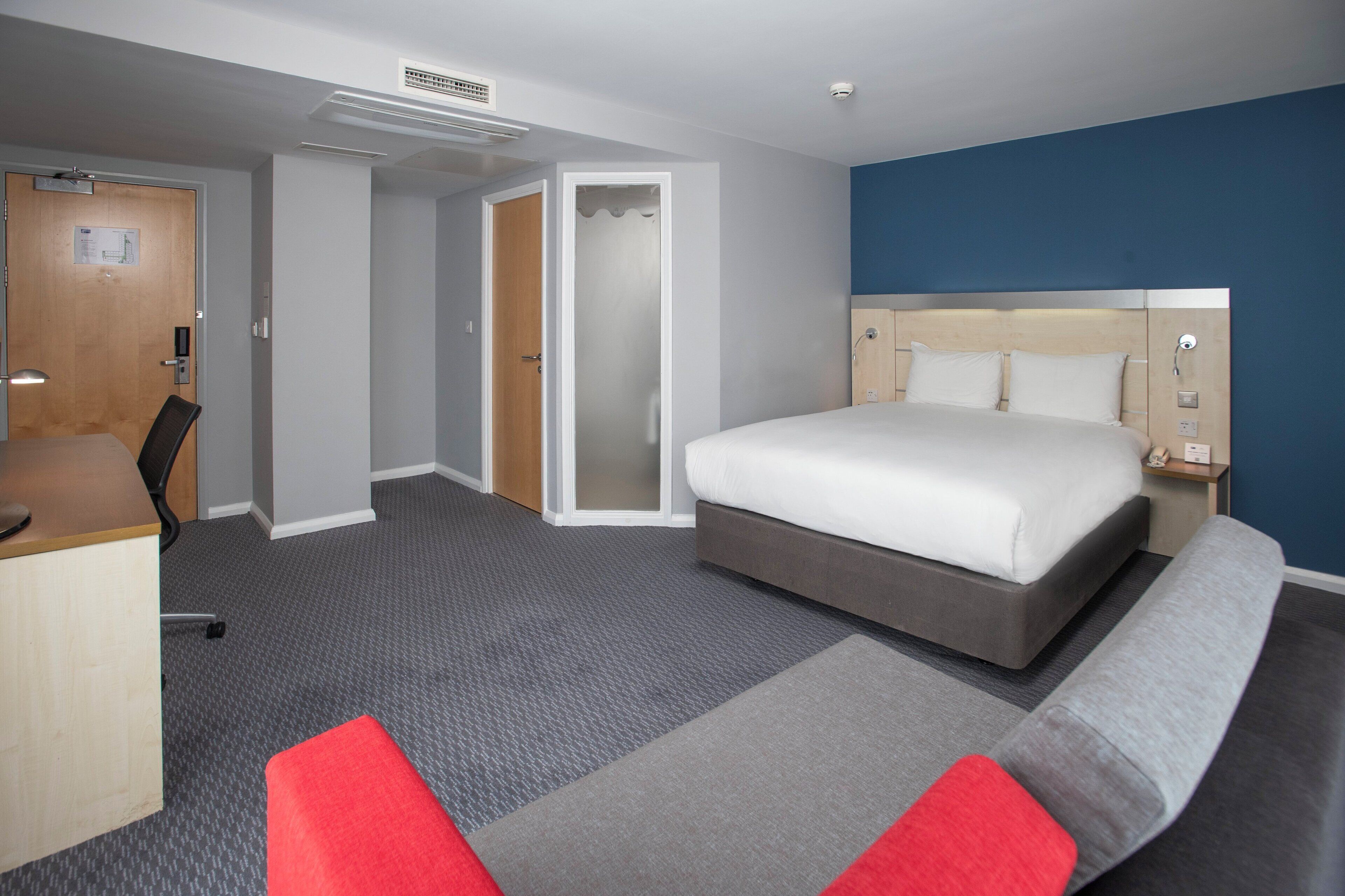 Una habitación de hotel con cama, escritorio, silla, sofá rojo y gris, y puerta al baño.
