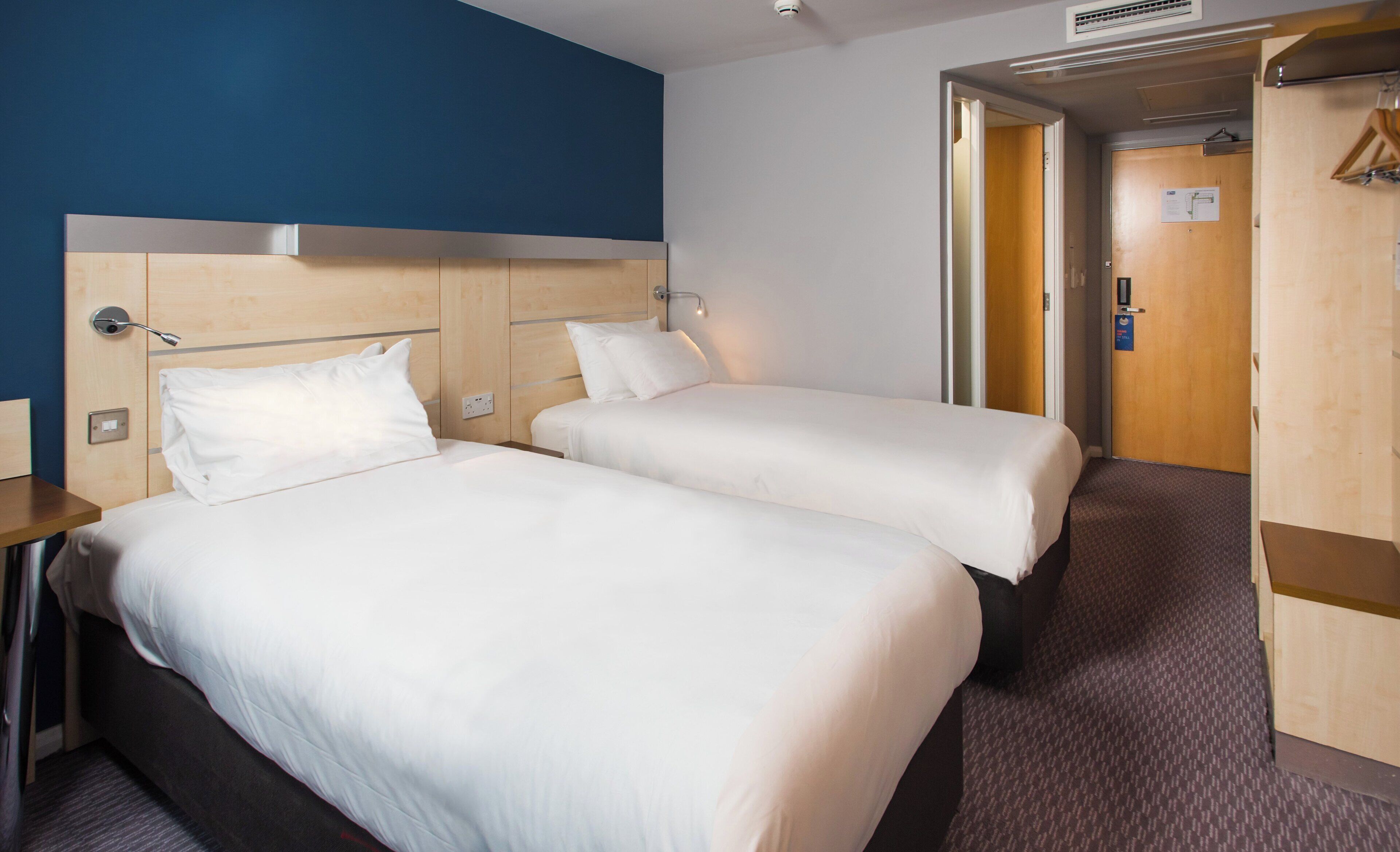 A hotel room with two beds, a wooden headboard, a bedside table, a wall socket, and a door leading to another room.
