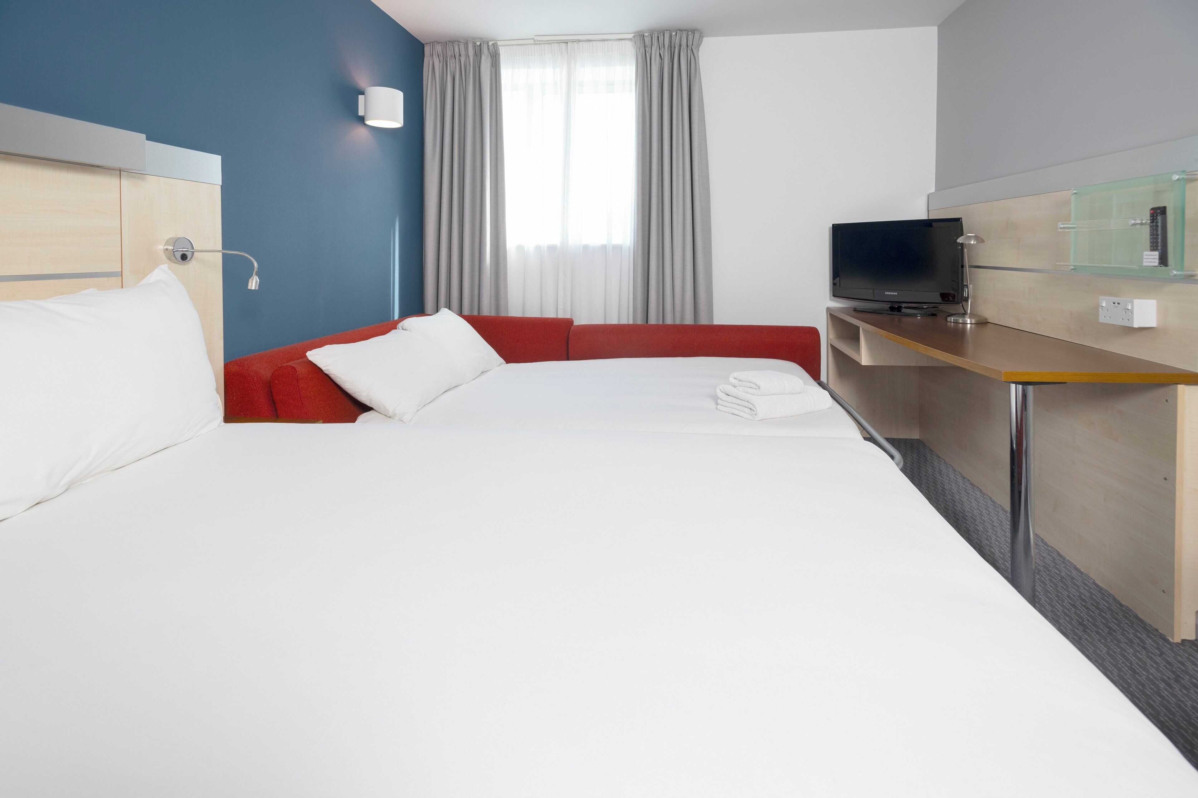 A hotel room with a large bed, a red sofa, a desk with a television, and a window with curtains.
