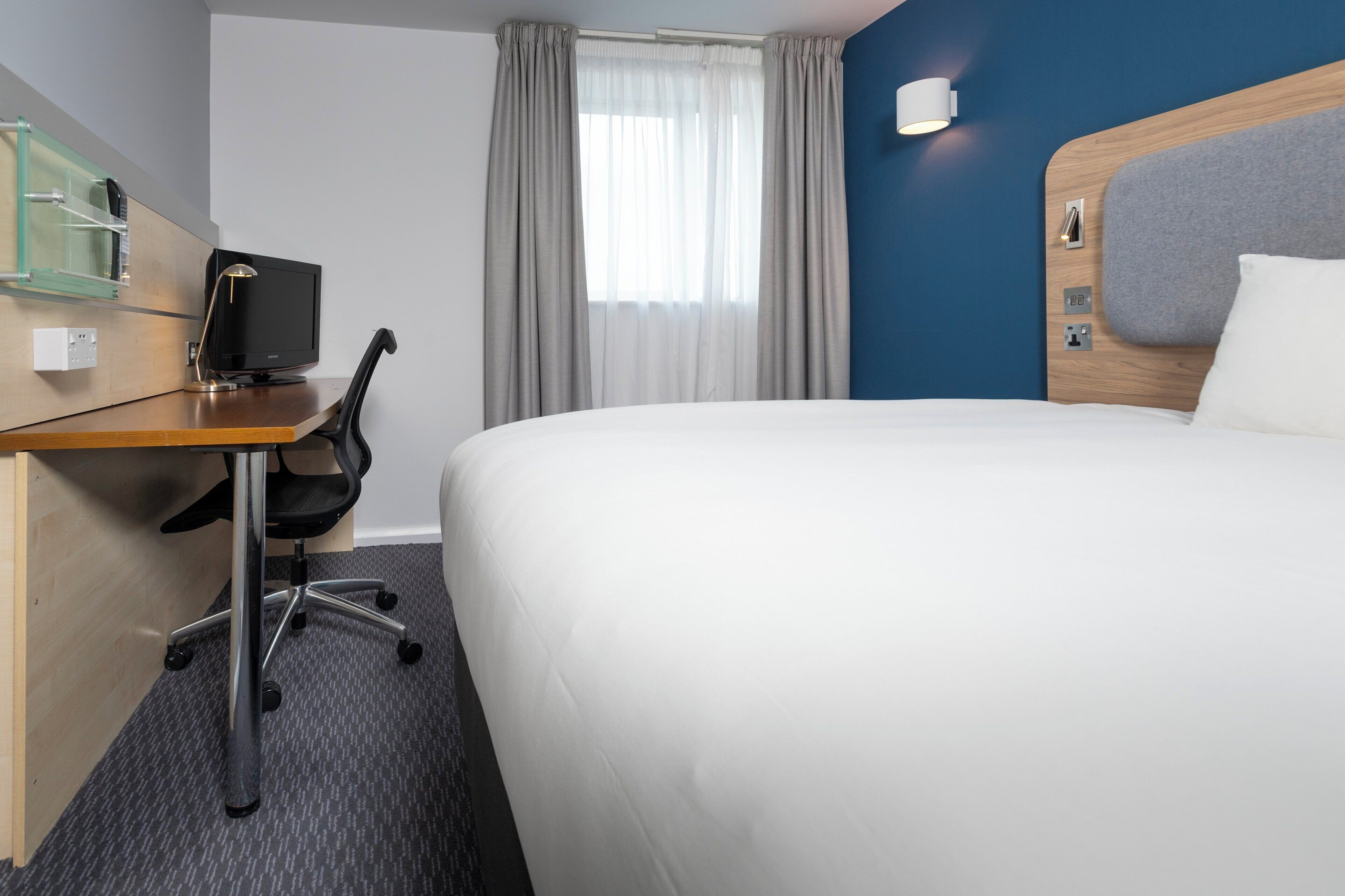 Habitación de hotel con cama, escritorio con computadora, silla y ventana con cortinas.