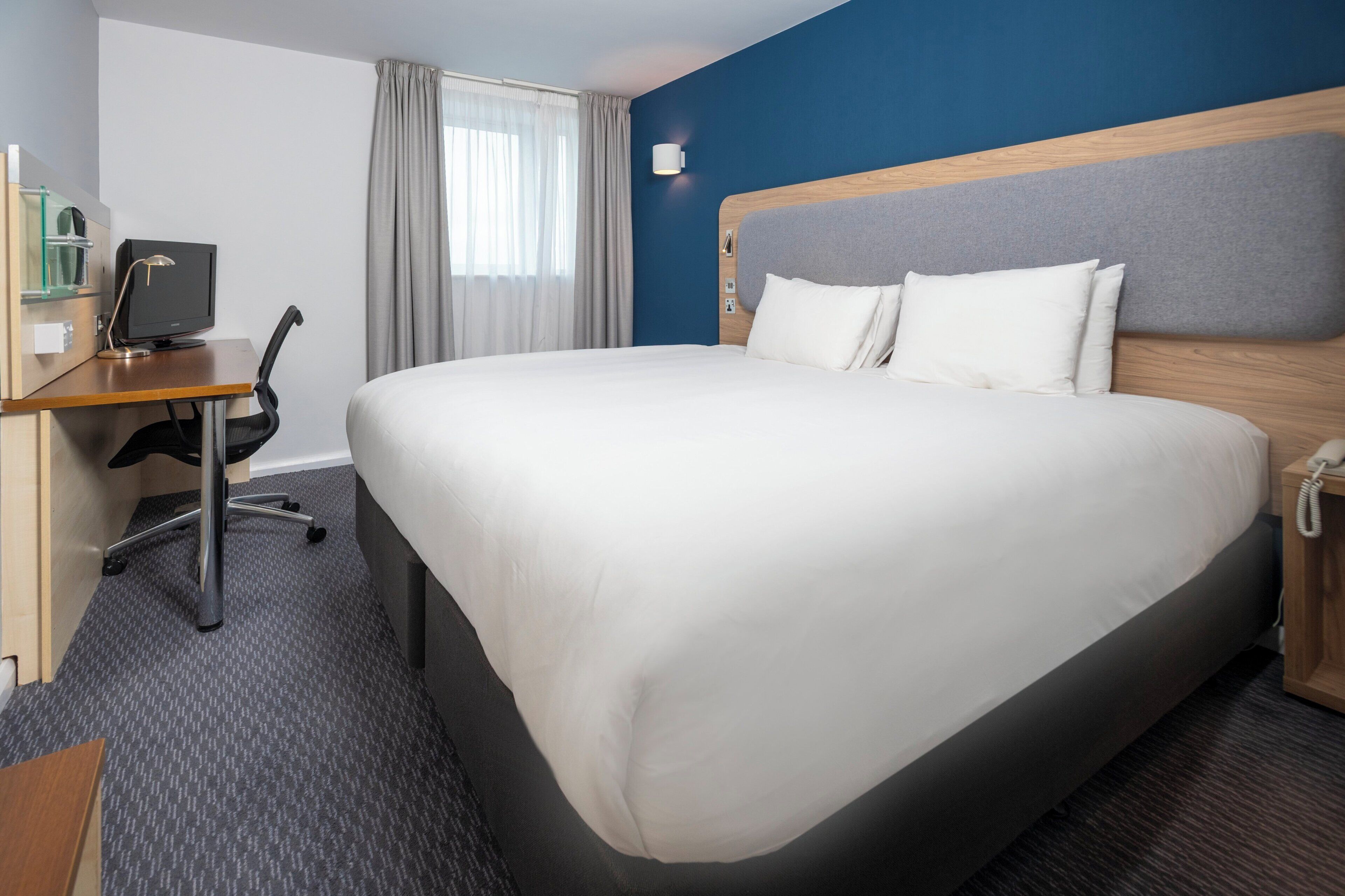 A hotel room with a large bed, a desk with a computer, a chair, and a telephone.