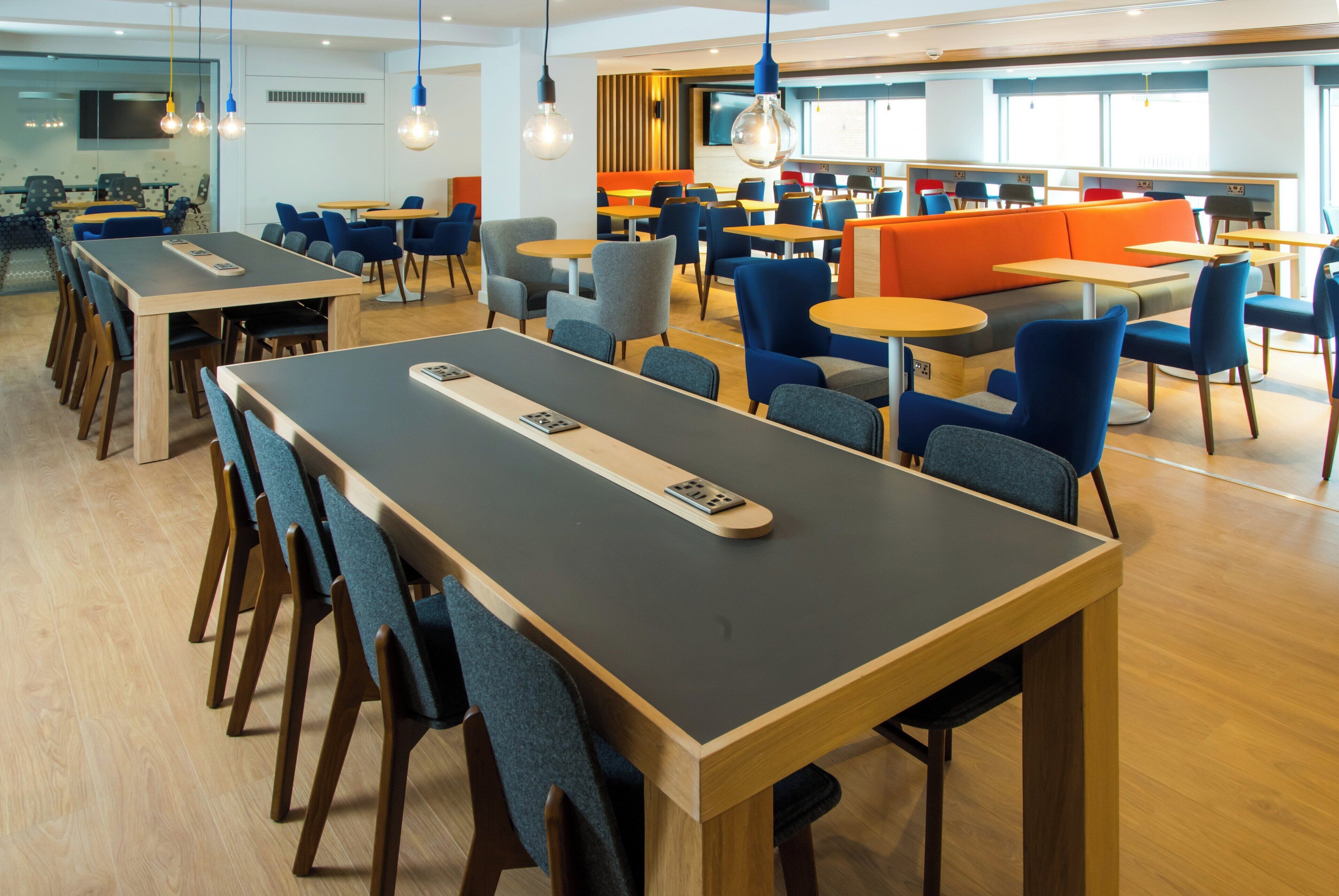 Un espace repas moderne doté de plusieurs tables et chaises, d’une longue table conviviale équipée d’une télécommande, et d’un coin salon avec des coussins orange.