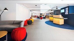 Egy modern szállodai előcsarnok, amelyben található egy recepciós pult, színes székekkel berendezett ülősarok, és a háttérben egy jól látható étterem.