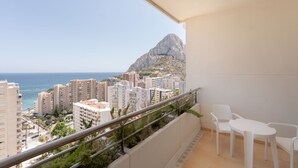 Un balcón con sillas y una mesa blancos, con vistas a una ciudad costera con edificios y una montaña.