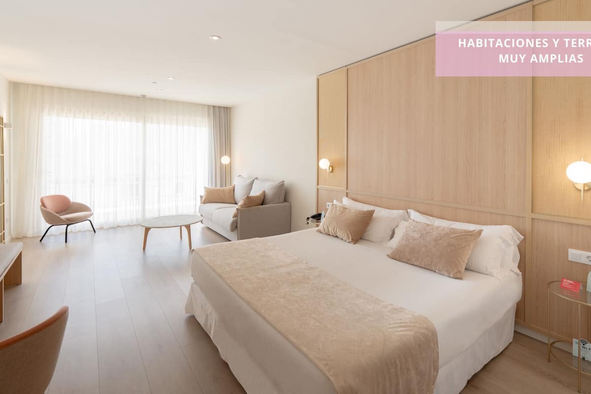 A spacious hotel room with a large bed, a sofa, a chair, and a small table. There are wall-mounted lights and a power outlet on the side table.