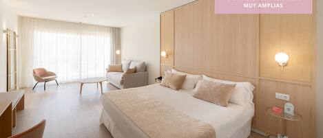 A spacious hotel room with a large bed, a sofa, a chair, and a small table. There are wall-mounted lights and a power outlet on the side table.