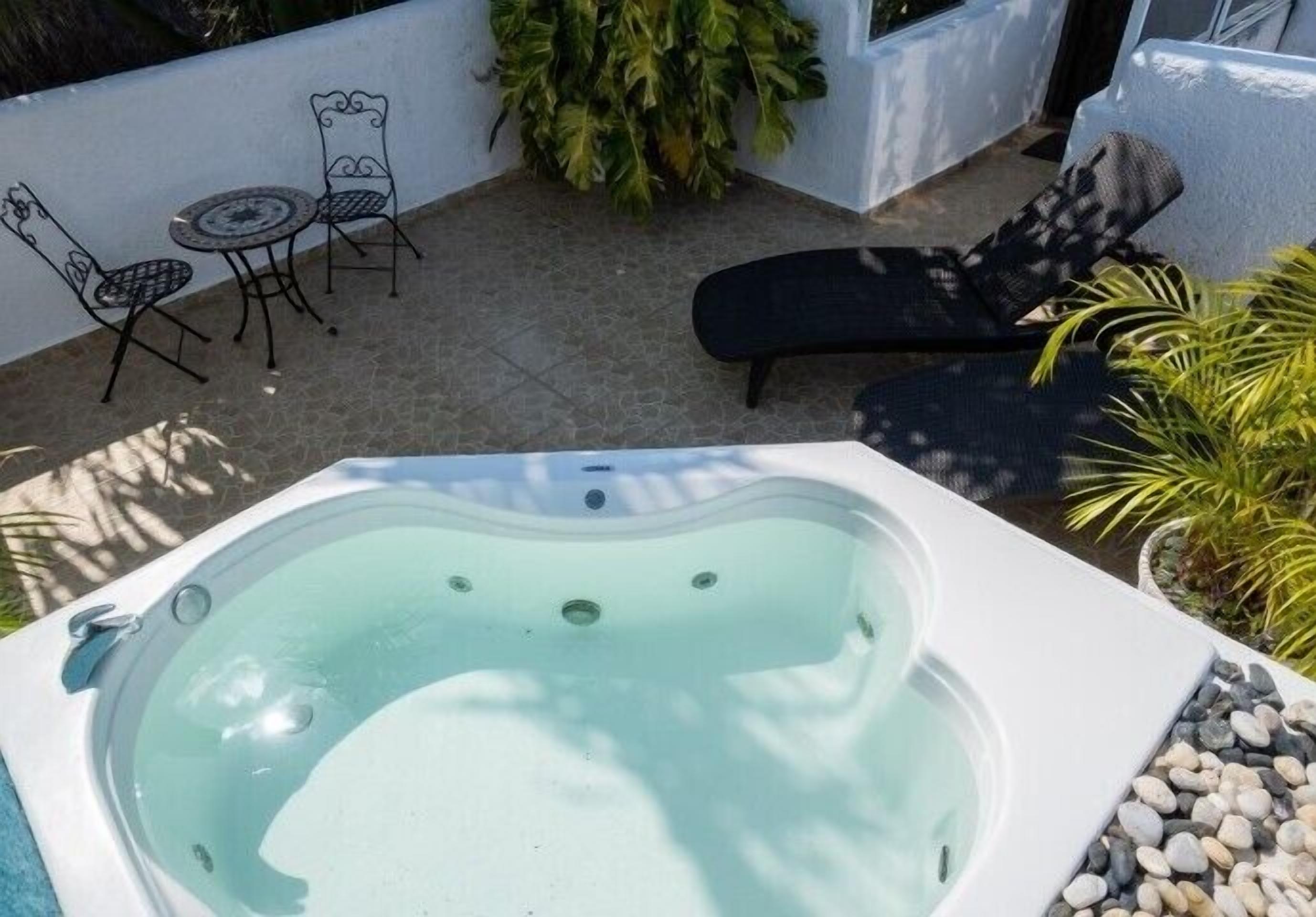 Ein Whirlpool in einer Terrassenanlage mit einem kleinen Tisch und Stühlen, umgeben von Pflanzen und einem Liegestuhl.