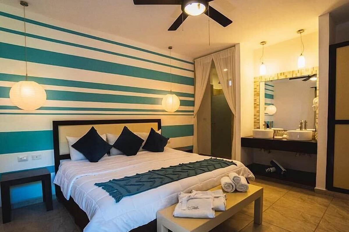A hotel room with a bed, bedside tables, a bench with towels, a mirror, and a bathroom with a sink and mirror.