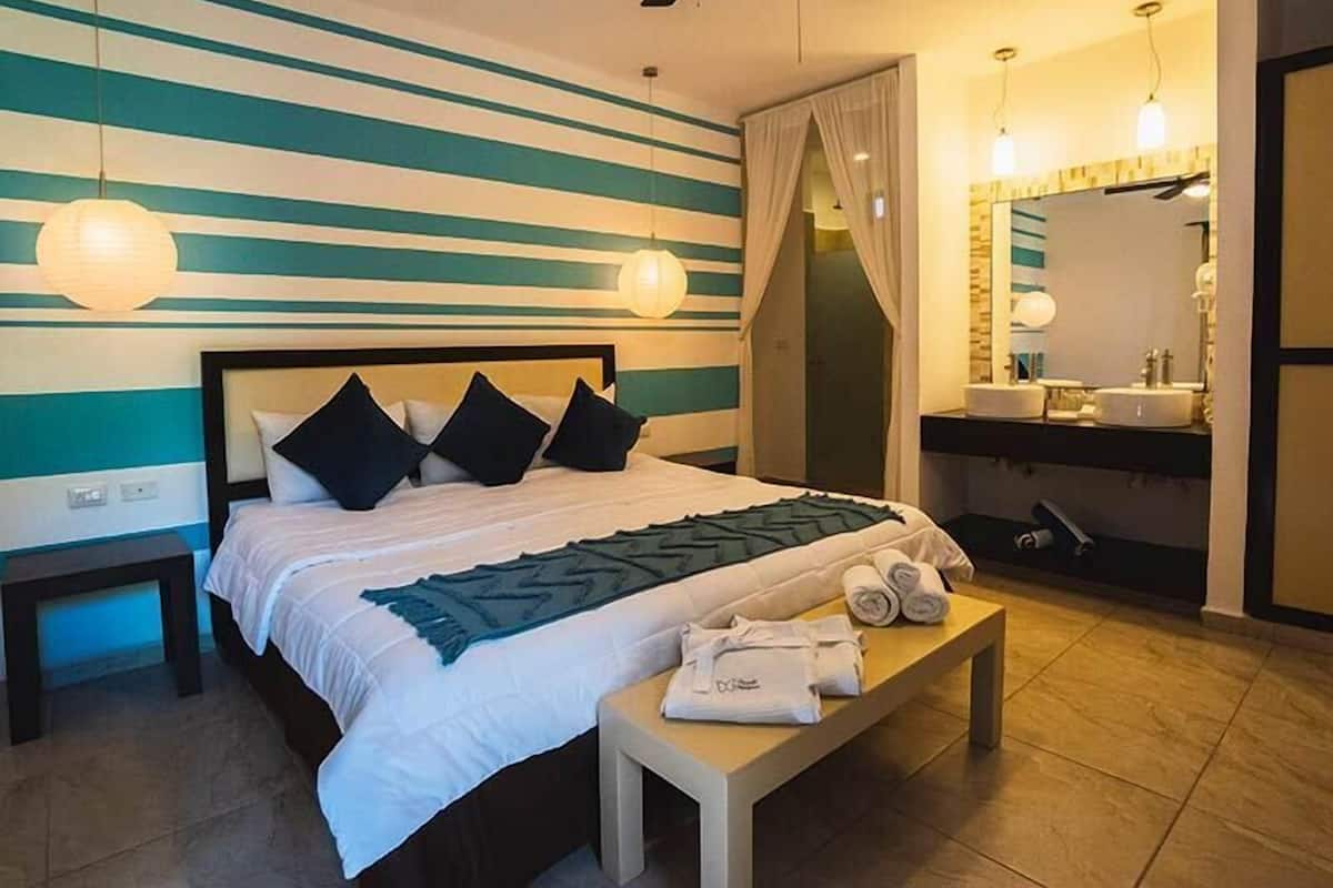 A hotel room with a bed, bedside table, bench, and a bathroom with a sink and mirror.