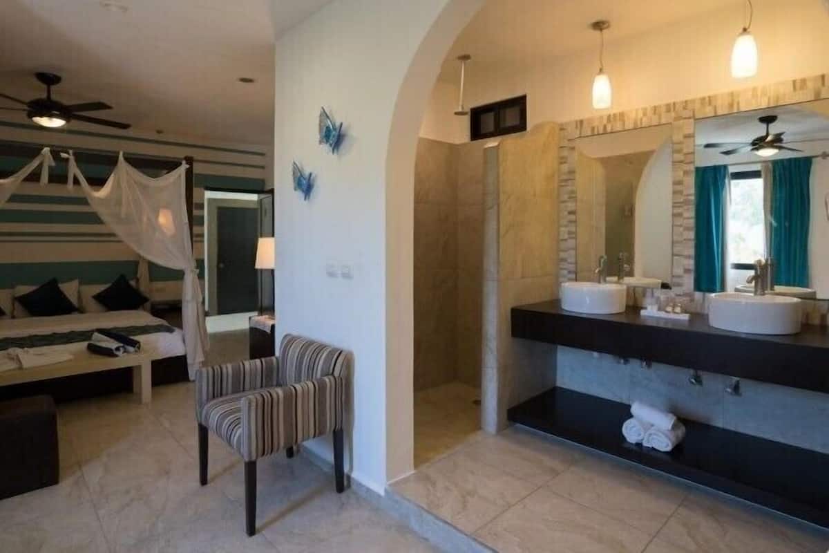 A modern hotel room with a bed, a chair, a shower area, and a large bathroom with a vanity and mirror.