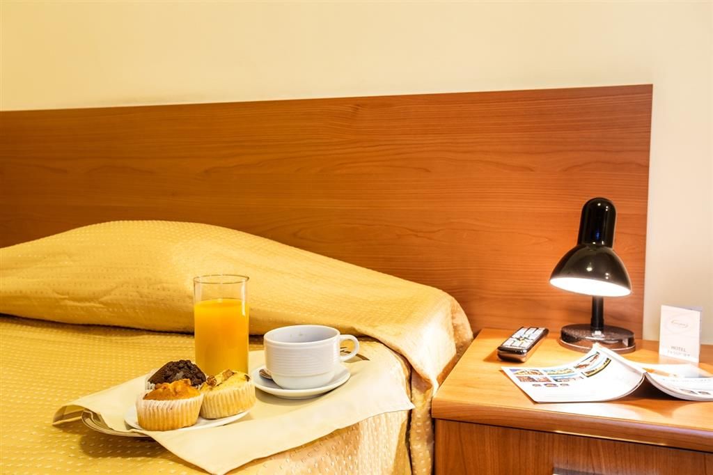 Una cama bien hecha con cabecera de madera, una mesita de noche con una lámpara, una taza de café, un vaso de jugo de naranja y un muffin en una bandeja.